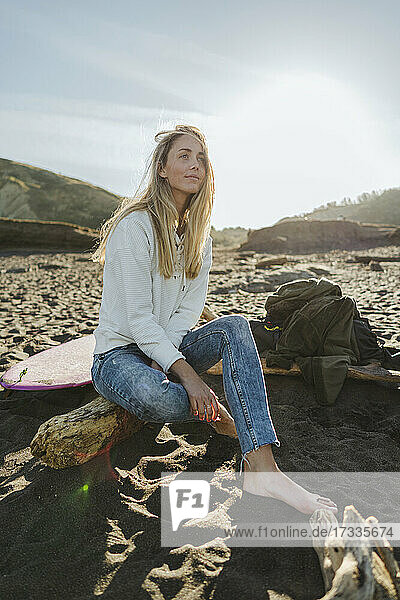 Schöne Frau mit blondem Haar sitzt auf einem Baumstamm am Strand