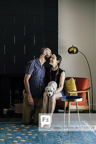 Kümmernder Mann küsst Frau mit Hund auf Stuhl im Wohnzimmer sitzend