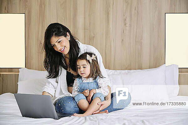 Lächelnde Mutter  die einen Laptop benutzt  während sie mit ihrer Tochter zu Hause auf dem Bett sitzt