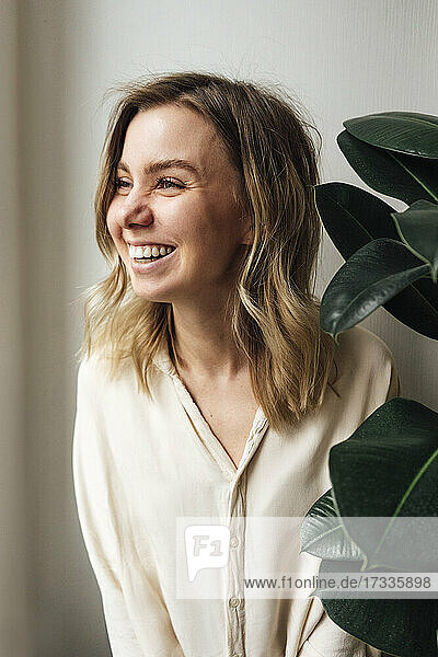 Cheerful blond woman looking away by plant at home