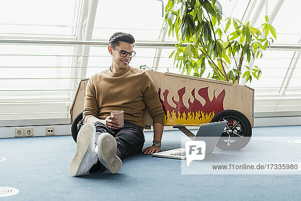 Male professional smiling while sitting by laptop on toy car in creative office