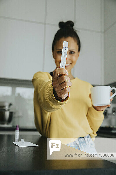 Lächelnde Frau zeigt Schnelltest-Kit und hält Becher in der Küche