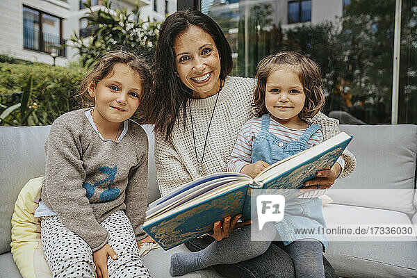 Lächelnde Mutter mit süßer Tochter  die ein Buch auf dem Sofa im Hinterhof hält