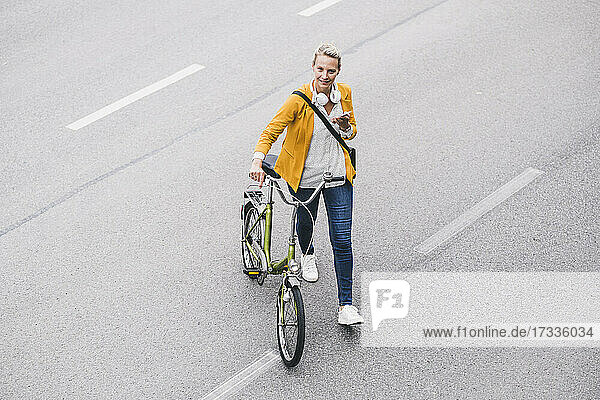 Geschäftsfrau mit Mobiltelefon in der Hand  während sie mit dem Fahrrad auf der Straße geht