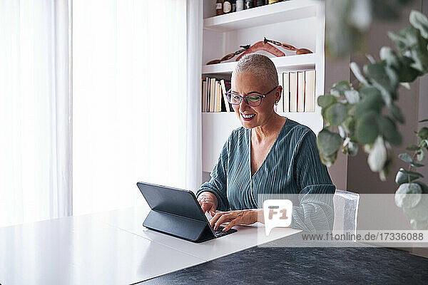 Lächelnde Frau mit rasiertem Kopf  die zu Hause ein digitales Tablet benutzt