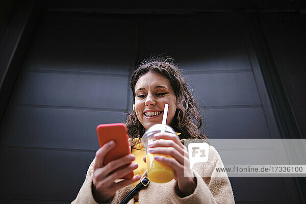 Lächelnde junge Frau mit Smoothie und Smartphone in der Nähe einer schwarzen Tür