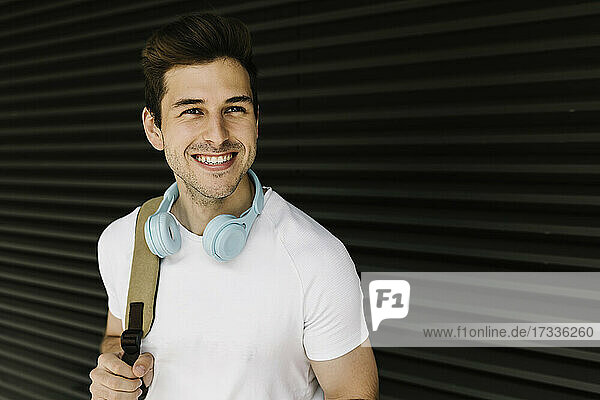 Lächelnder junger Mann schaut weg  während er einen Rucksack vor einem Fensterladen trägt