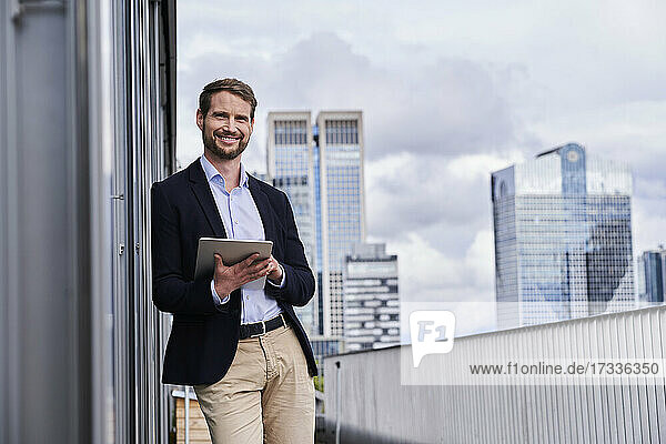 Smiling businessman holding digital tablet leaning on building terrace