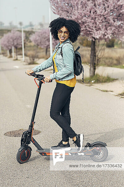 Lächelnde junge Frau steht mit elektrischem Roller auf der Straße