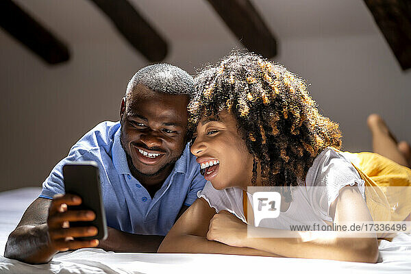 Lächelndes Paar  das ein Smartphone benutzt  während es zu Hause auf dem Bett liegt