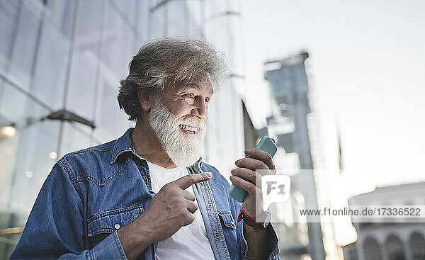 Happy white bearded mature man pointing at mobile phone in front of building