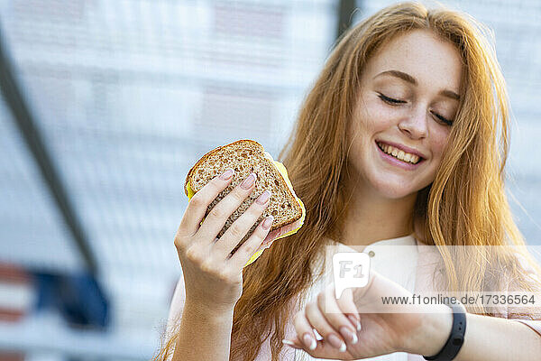 Lächelnde weibliche Fachkraft  die die Zeit überprüft  während sie ein Sandwich am Bahnhof isst