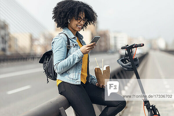 Lächelnde Frau  die ein Smartphone benutzt und Essen in der Hand hält  während sie sich auf ein Geländer stützt