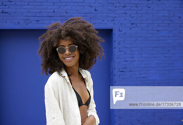 Lächelnde junge Afro-Frau mit Sonnenbrille vor einer blauen Wand