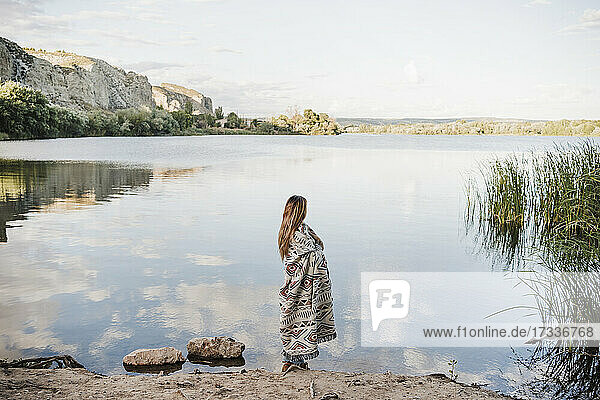 Junge Frau in Decke eingewickelt am Seeufer stehend