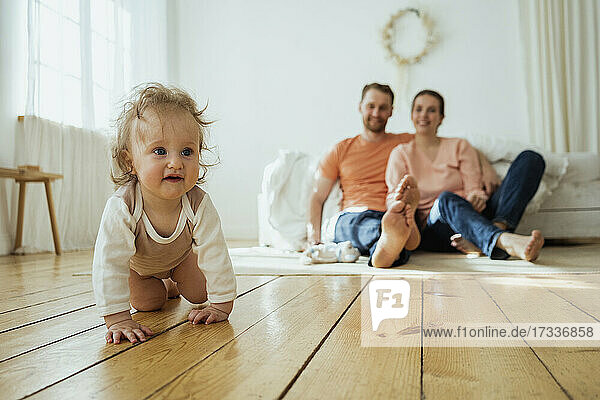 Niedliches Mädchen krabbelt  während Mutter und Vater zu Hause auf dem Boden sitzen