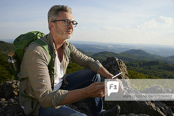 Mann hält digitales Tablet  während er auf einem Berg sitzt