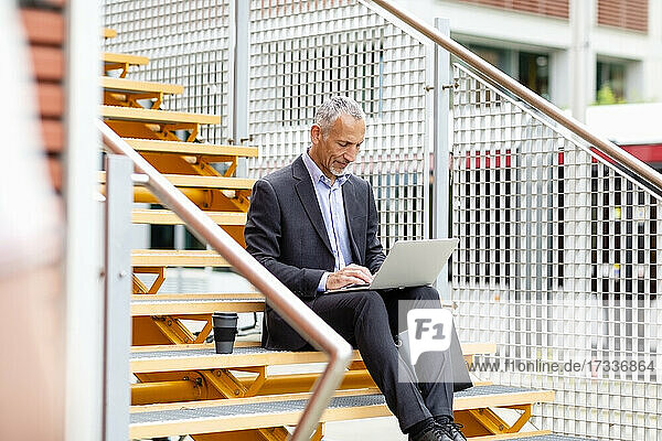 Männlicher Berufstätiger in Geschäftskleidung  der auf einer Treppe sitzend am Laptop arbeitet