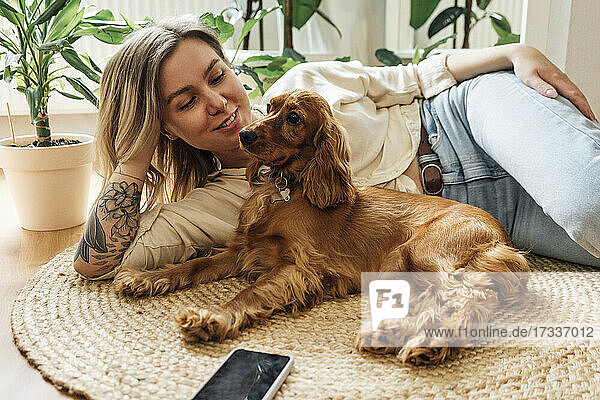 Beautiful woman looking at dog while relaxing on rug in living room