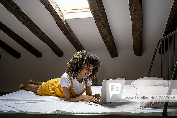 Lächelnde junge Frau  die einen Laptop benutzt  während sie auf einem Bett auf dem Dachboden liegt