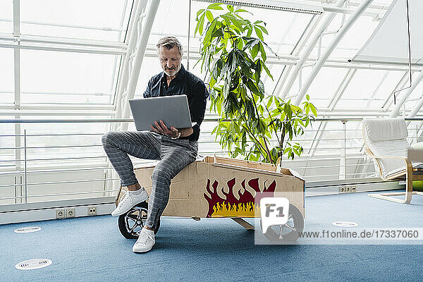 Geschäftsmann  der einen Laptop benutzt und auf einem Spielzeugauto im Kreativbüro sitzt