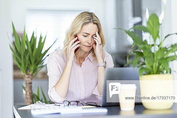 Müde weibliche Fachkraft mit Kopf in der Hand  die im Büro zu Hause mit einem Smartphone spricht