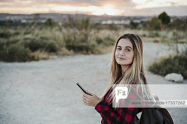 Beautiful young woman wearing backpack holding smart phone during sunset