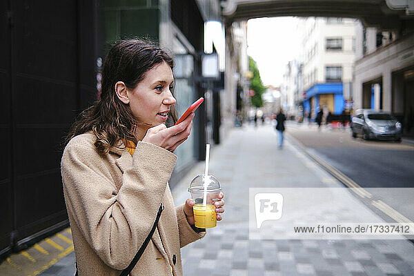 Junge Frau hält einen Smoothie in der Hand und sendet eine Sprachnachricht über ihr Smartphone auf dem Fußweg