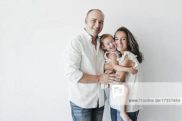 Happy woman carrying daughter in front of white background standing next to husband