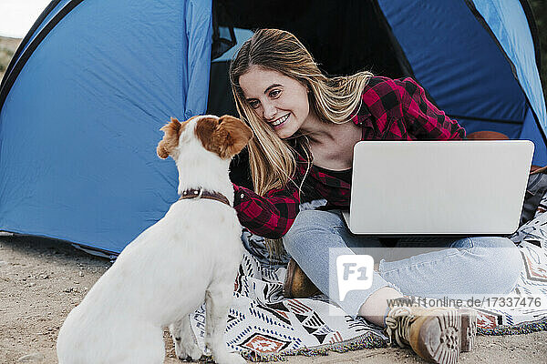 Happy young digital nomad stroking dog while sitting with laptop outside tent