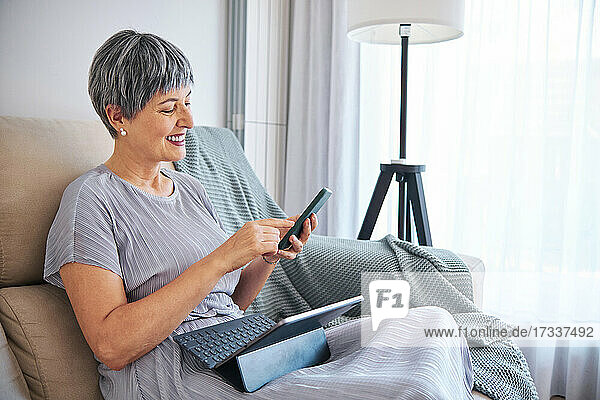 Frau mit digitalem Tablet sitzend  während sie zu Hause ihr Smartphone benutzt