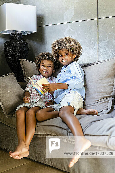Afro brothers with book sitting on sofa at home