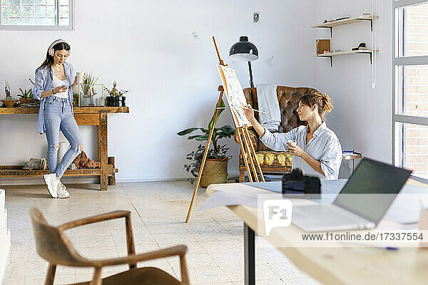 Female artist painting while photographer colleague listening music in studio