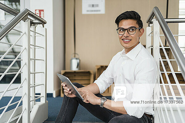 Männlicher Fachmann lächelt  während er mit einem digitalen Tablet an einem Geländer sitzt
