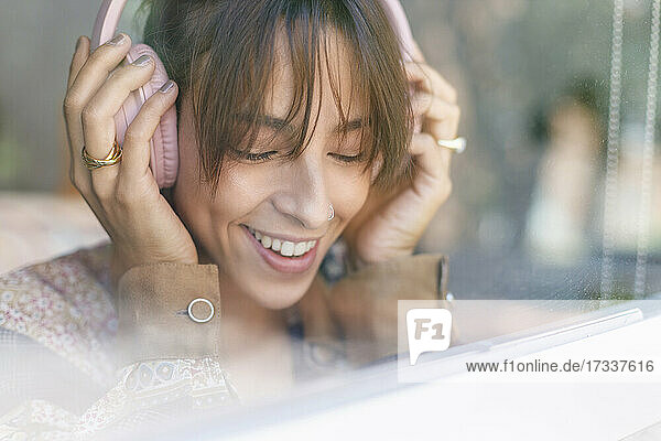Lächelnde Frau  die über Kopfhörer Musik hört  gesehen durch ein Fenster