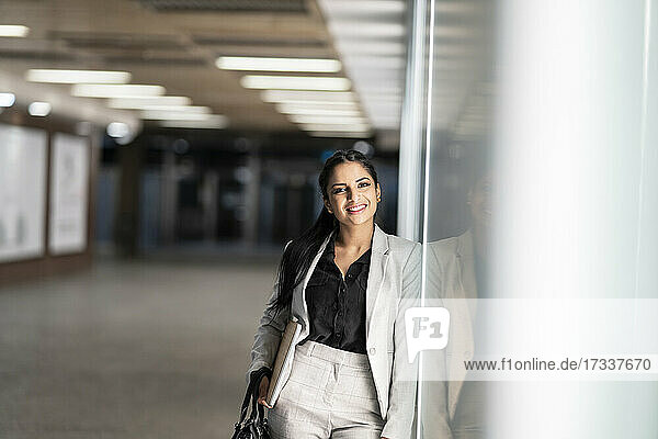 Smiling businesswoman with laptop and purse leaning on glass window