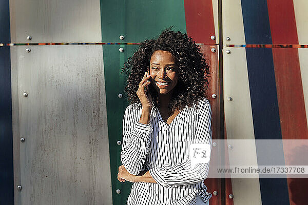 Smiling curly haired woman talking on smart phone while standing in front of wall looking away