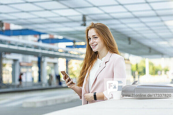 Junge Geschäftsfrau schaut weg  während sie ihr Smartphone am Bahnhof hält