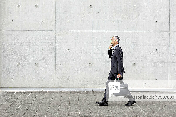 Männlicher Berufstätiger mit Aktentasche beim Gehen  während er mit seinem Smartphone telefoniert