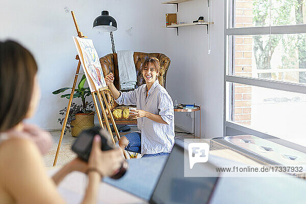 Smiling female painter looking at photographer colleague holding camera at studio
