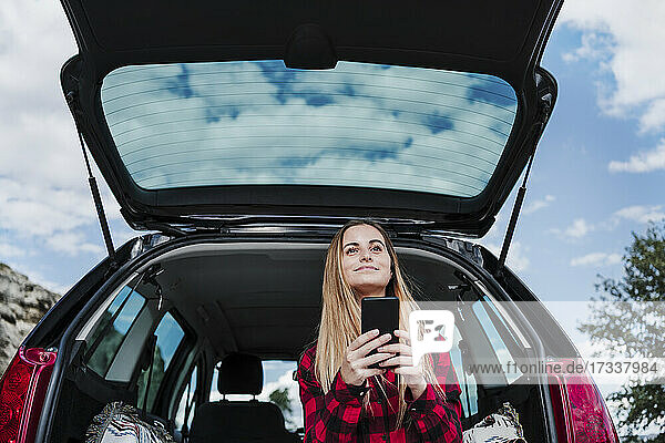 Blonde Frau sitzt mit Smartphone im Kofferraum eines Autos