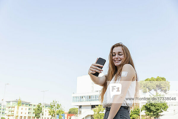 Lächelnde Geschäftsfrau  die ein Selfie mit ihrem Mobiltelefon macht  nachdem sie sich vor dem Himmel geimpft hat