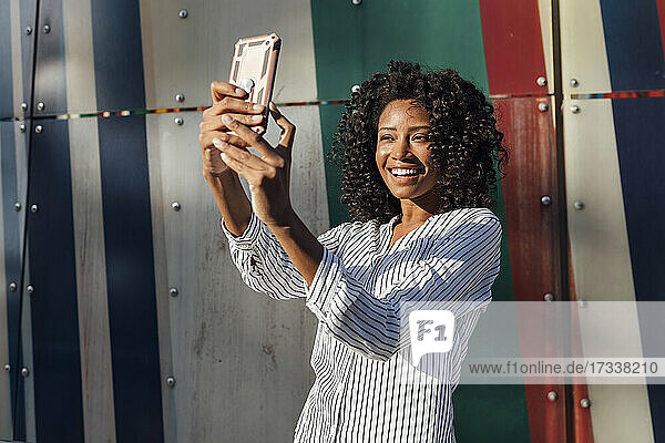 Happy young woman taking selfie through smart phone while standing in front of wall