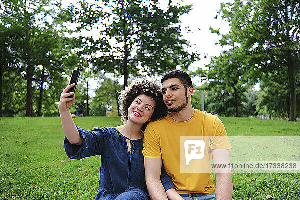 Lächelnde junge Frau  die ein Selfie mit ihrem Freund über ihr Smartphone macht  während sie im Park sitzt