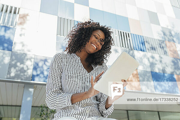 Fröhliche  lockig behaarte Geschäftsfrau  die vor einem Bürogebäude sitzt und ein digitales Tablet benutzt