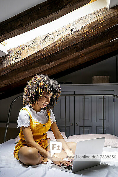 Afro-Frau  die einen Laptop benutzt  während sie auf dem Dachboden zu Hause auf dem Bett sitzt