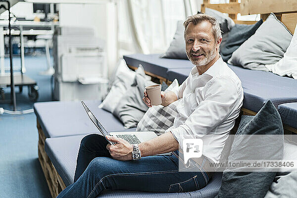 Smiling male professional with disposable cup and laptop sitting on sofa