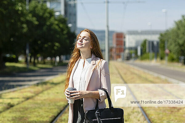 Redhead businesswoman with eyes closed standing at railroad track during sunny day