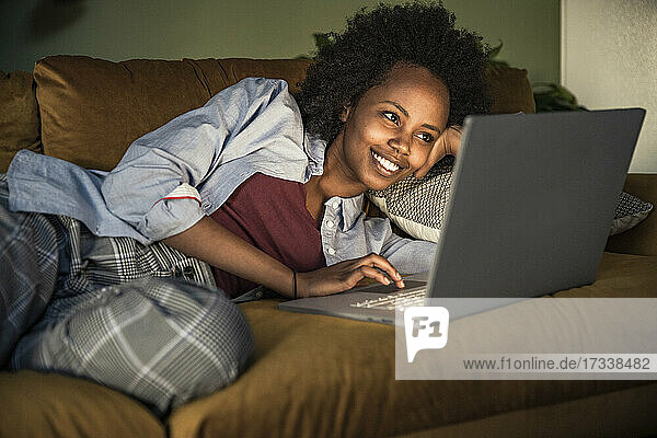 Junge Frau benutzt einen Laptop  während sie zu Hause auf dem Sofa liegt