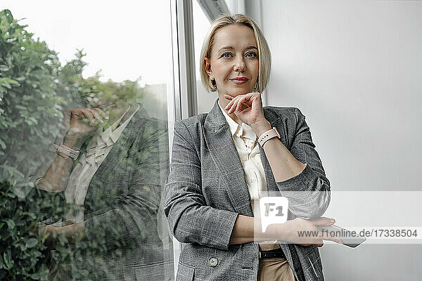 Schöne blonde Geschäftsfrau stehend mit Hand auf Kinn durch Glasfenster im Büro